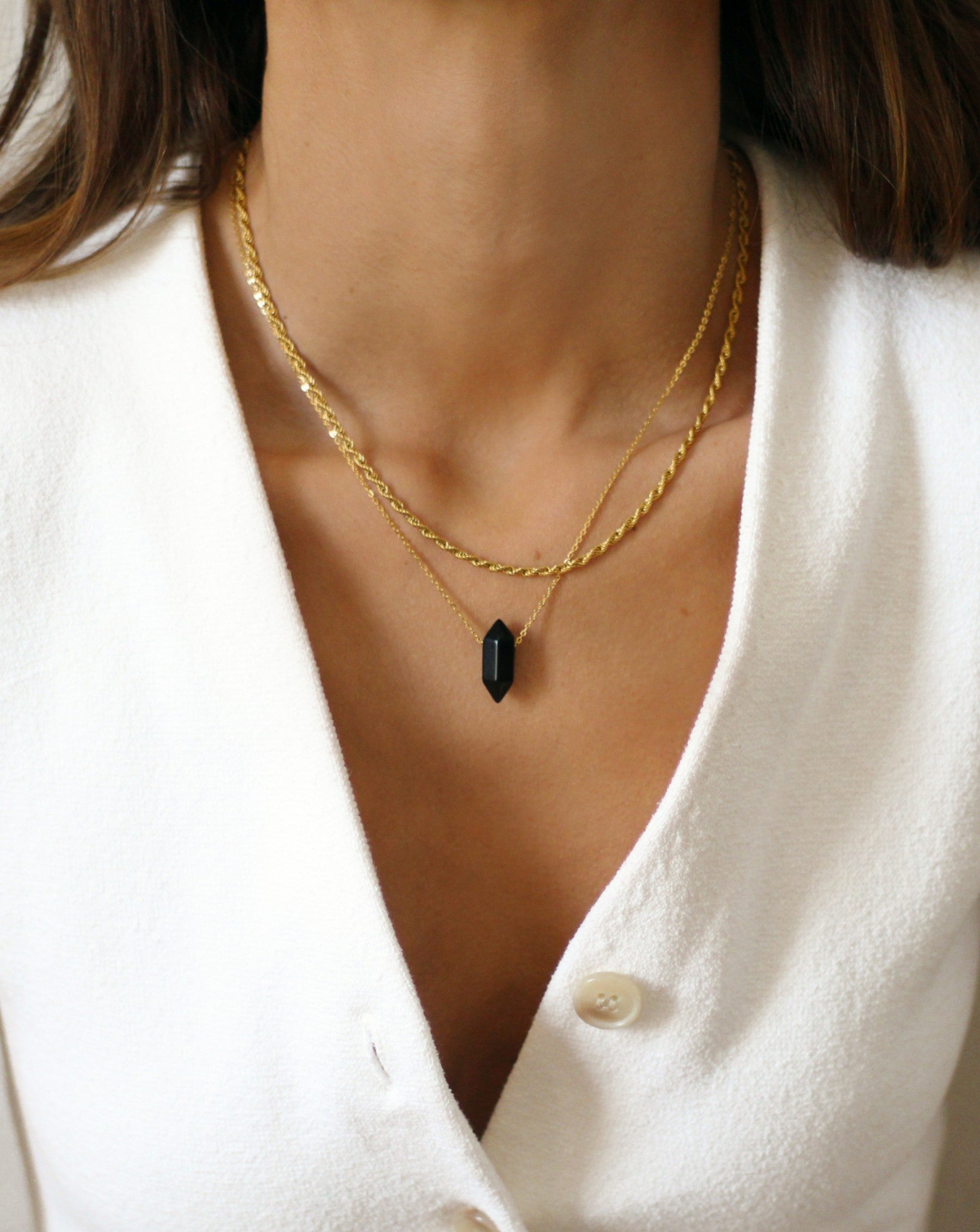 A person wearing a gold rope necklace with a white sweater and a neutral facial expression.