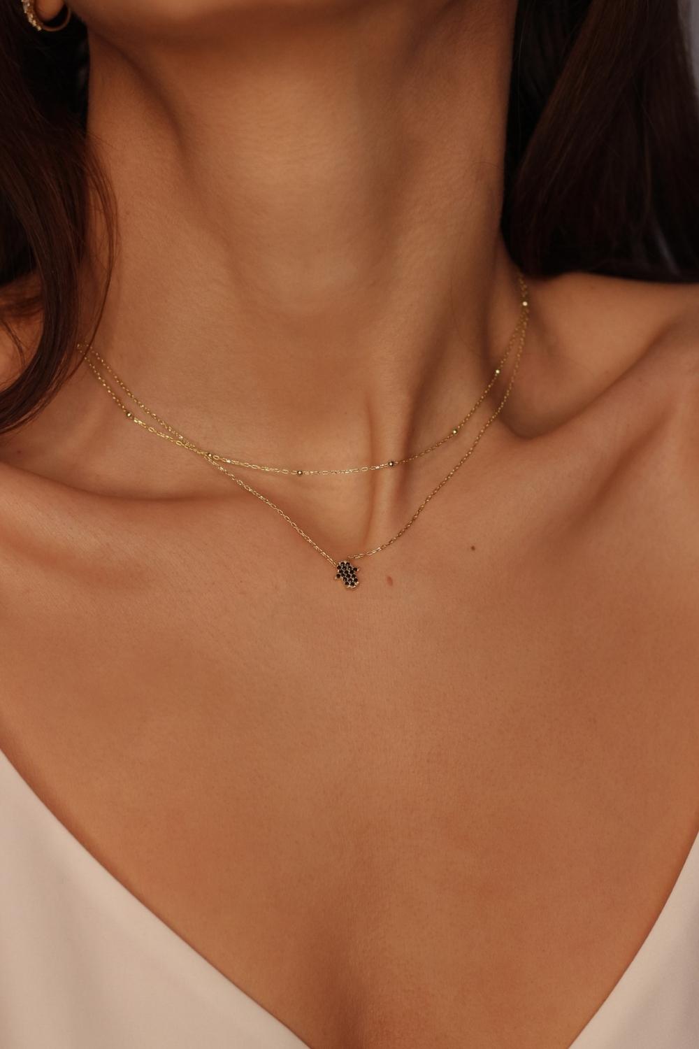 a black Hamsa hand charm necklace in 18k gold plated silver. The necklace features a delicate chain with a beautifully designed Hamsa hand charm pendant. The Hamsa hand symbol is believed to bring protection, good luck, and ward off evil. The necklace is designed to be worn comfortably and can be adjusted to fit different neck sizes.