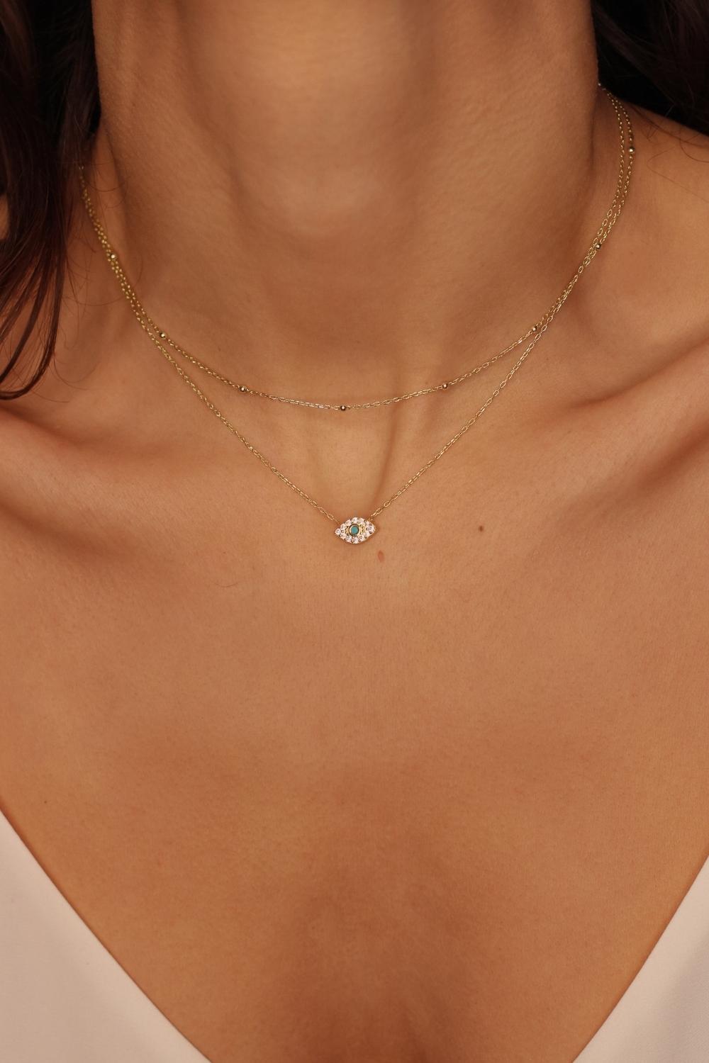 A layered necklaces featuring an evil eye charm necklace in gold and a satellite chain choker. The set includes a delicate 18k gold plated chain with an intricately designed evil eye charm pendant. Alongside the evil eye charm necklace, a satellite chain choker is also included, adding a trendy and minimalist touch. The satellite chain choker features small, evenly spaced interlinked gold beads, creating a delicate and stylish look.