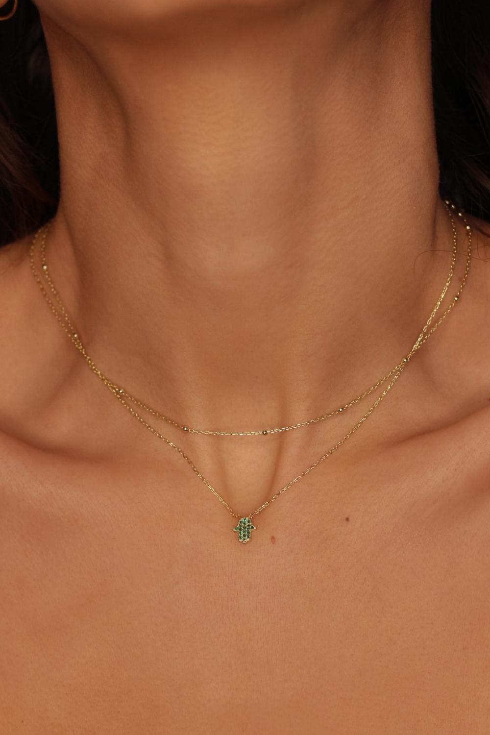 A Green Hamsa hand charm necklace in 18k gold plated silver. The necklace features a delicate chain with a beautifully designed Hamsa hand charm pendant. The Hamsa hand symbol is believed to bring protection, good luck, and ward off evil. The necklace is designed to be worn comfortably and can be adjusted to fit different neck sizes.