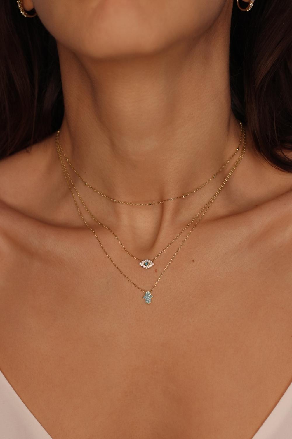 A blue Hamsa hand charm necklace in 18k gold plated silver. The necklace features a delicate chain with a beautifully designed Hamsa hand charm pendant. The Hamsa hand symbol is believed to bring protection, good luck, and ward off evil. The necklace is designed to be worn comfortably and can be adjusted to fit different neck sizes.