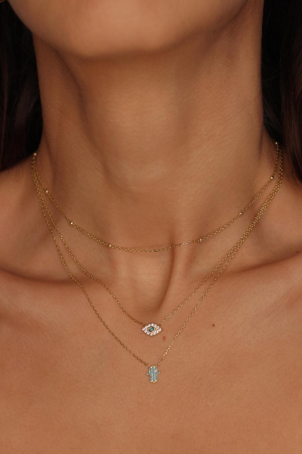 A layered necklaces featuring an evil eye charm necklace, hamsa necklace in gold and a satellite chain choker. The set includes a delicate 18k gold plated chain with an intricately designed evil eye charm pendant. Alongside the evil eye charm necklace, a satellite chain choker is also included, adding a trendy and minimalist touch. The satellite chain choker features small, evenly spaced interlinked gold beads, creating a delicate and stylish look.