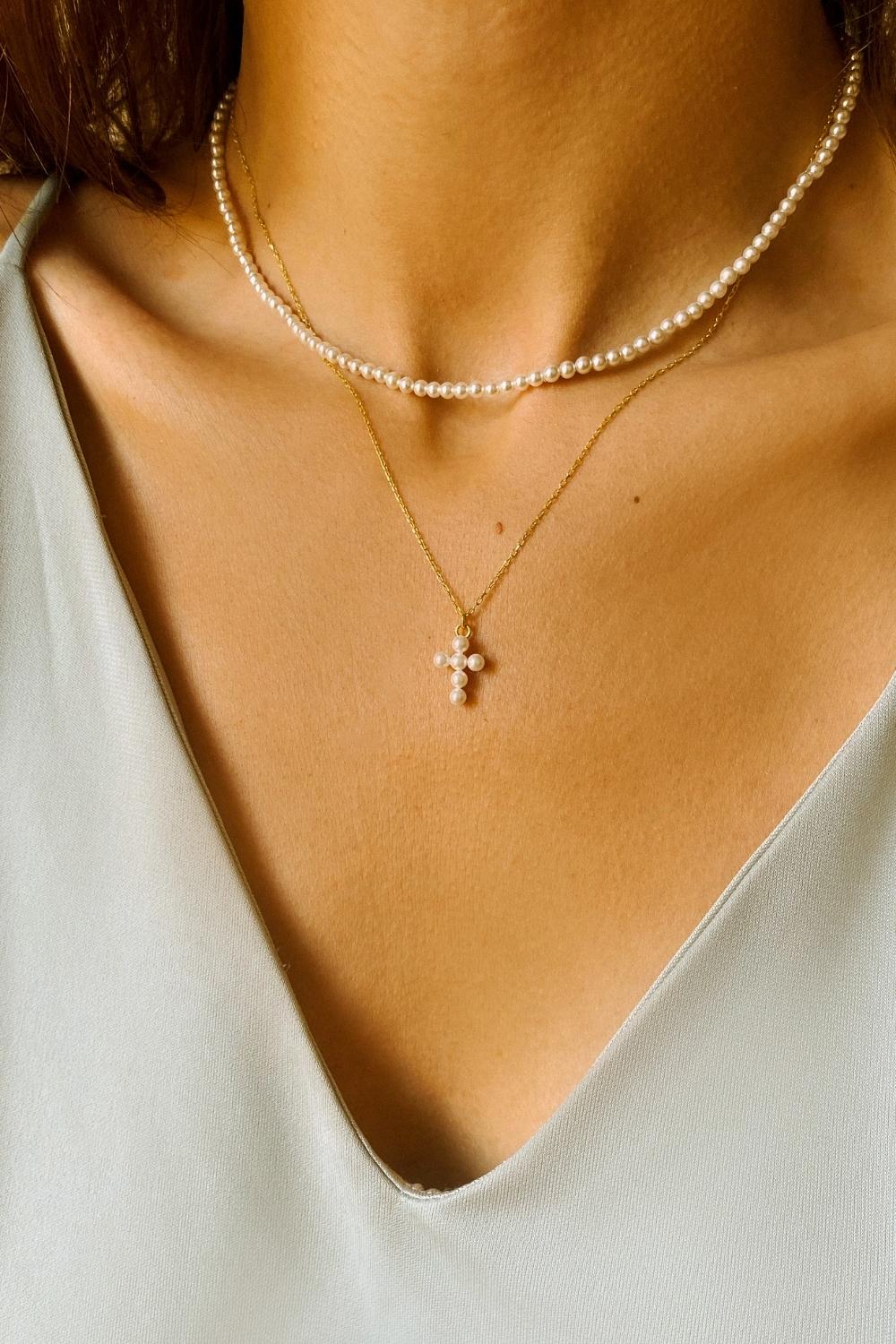 A layered pearl necklace set, featuring a pearl cross pendant necklace and a beaded pearl choker. The pearl cross pendant necklace is crafted from delicate gold-plated silver and features a cross pendant adorned with lustrous white pearls. The necklace is layered with a beaded pearl choker, which features delicate gold chains and lustrous white pearls threaded onto the chains.