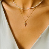 A layered pearl necklace set, featuring a pearl cross pendant necklace and a beaded pearl choker. The pearl cross pendant necklace is crafted from delicate gold-plated silver and features a cross pendant adorned with lustrous white pearls. The necklace is layered with a beaded pearl choker, which features delicate gold chains and lustrous white pearls threaded onto the chains.
