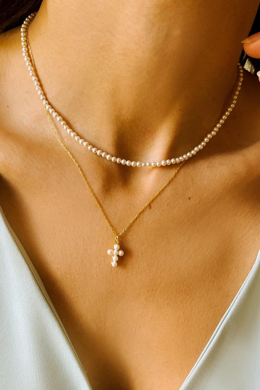 A layered pearl necklace set, featuring a pearl cross pendant necklace and a beaded pearl choker. The pearl cross pendant necklace is crafted from delicate gold-plated silver and features a cross pendant adorned with lustrous white pearls. The necklace is layered with a beaded pearl choker, which features delicate gold chains and lustrous white pearls threaded onto the chains.