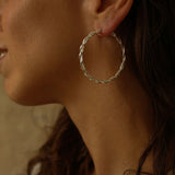a pair of silver hoop earrings, featuring a twisted design. The hoops are large in size, yet lightweight, making them comfortable to wear. The silver has a polished finish, reflecting light and enhancing their shine. These striking earrings add a bold and elegant touch to any outfit.