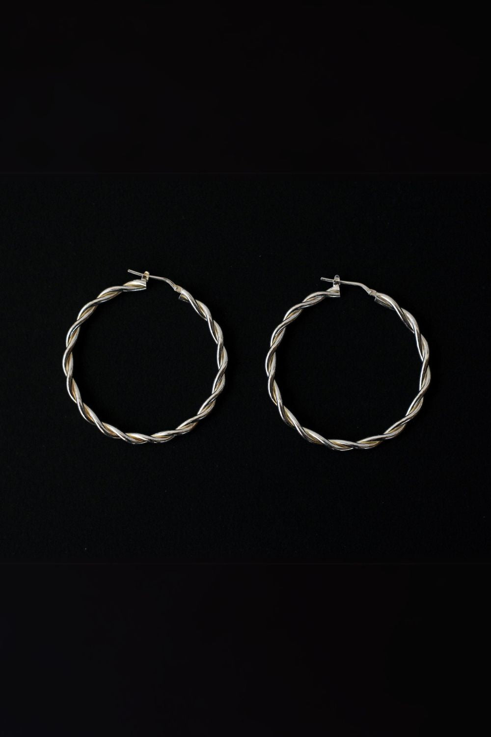 a pair of silver hoop earrings, featuring a twisted design. The hoops are large in size, yet lightweight, making them comfortable to wear. The silver has a polished finish, reflecting light and enhancing their shine. These striking earrings add a bold and elegant touch to any outfit.
