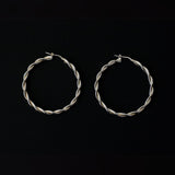 a pair of silver hoop earrings, featuring a twisted design. The hoops are large in size, yet lightweight, making them comfortable to wear. The silver has a polished finish, reflecting light and enhancing their shine. These striking earrings add a bold and elegant touch to any outfit.