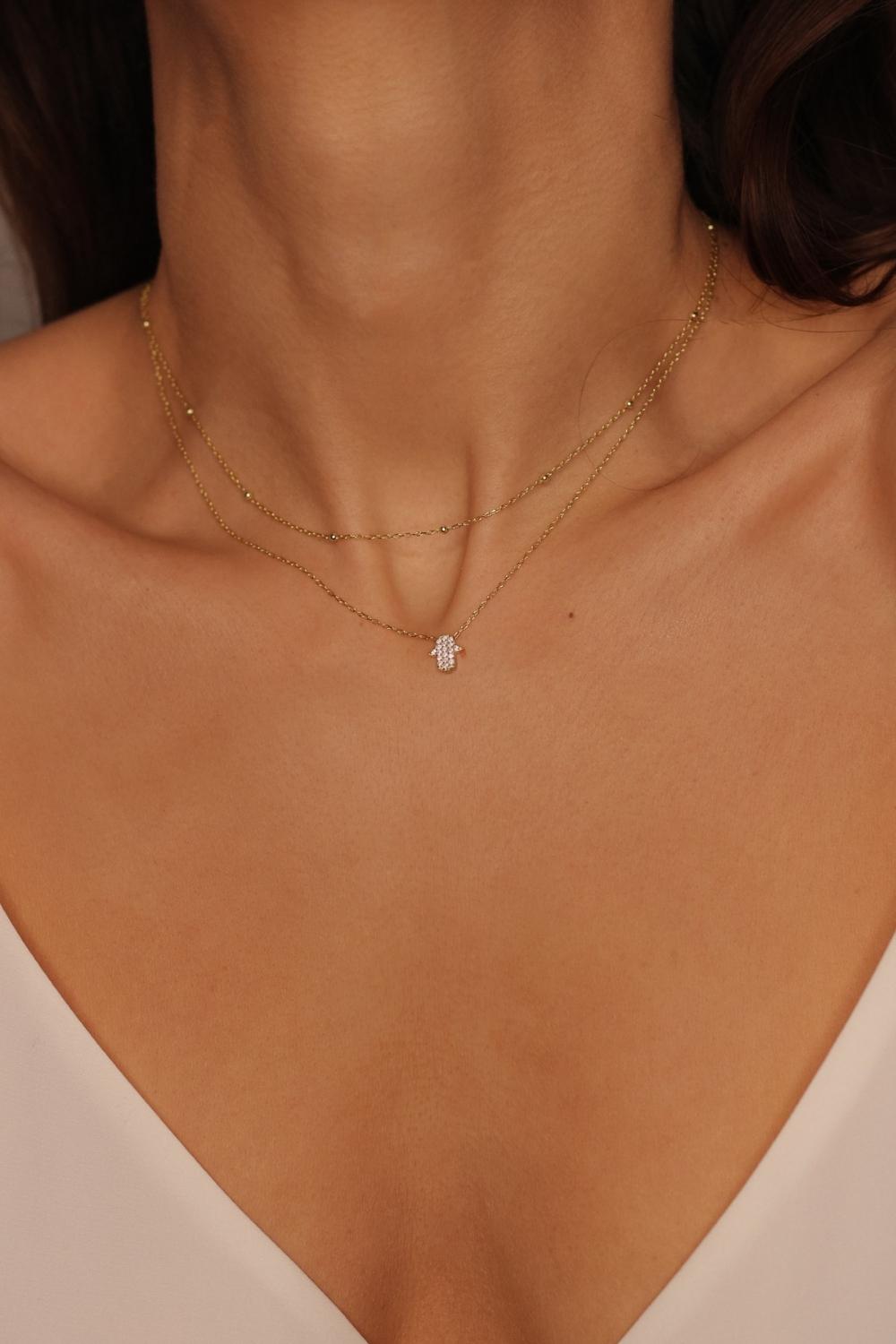 A White Hamsa hand charm necklace in 18k gold plated silver. The necklace features a delicate chain with a beautifully designed Hamsa hand charm pendant. The Hamsa hand symbol is believed to bring protection, good luck, and ward off evil. The necklace is designed to be worn comfortably and can be adjusted to fit different neck sizes.