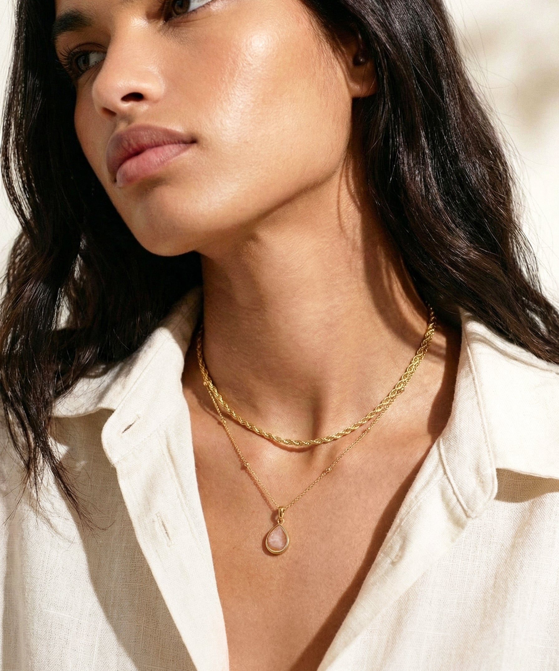 Woman wearing a gold necklace with a rose quartz pendant, looking off to the side.