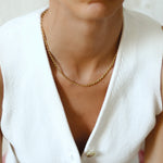 A person wearing a gold rope necklace with a white sweater and a neutral facial expression.