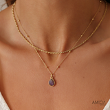 Gold necklace with a teardrop pendant on a person's neck, brand 'AMICA' visible.