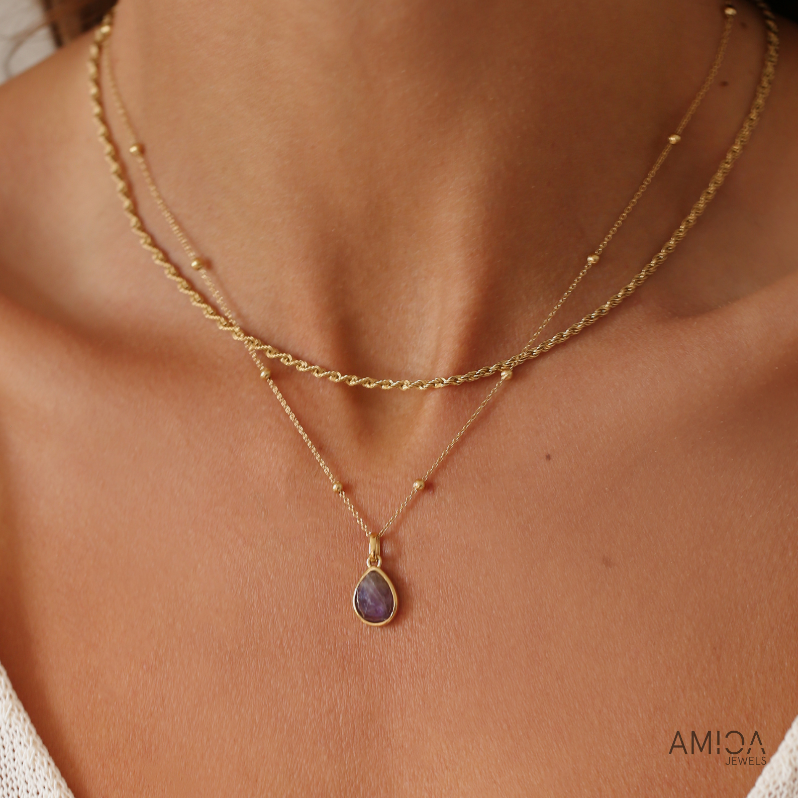 Gold necklace with a teardrop pendant on a person's neck, brand 'AMICA' visible.