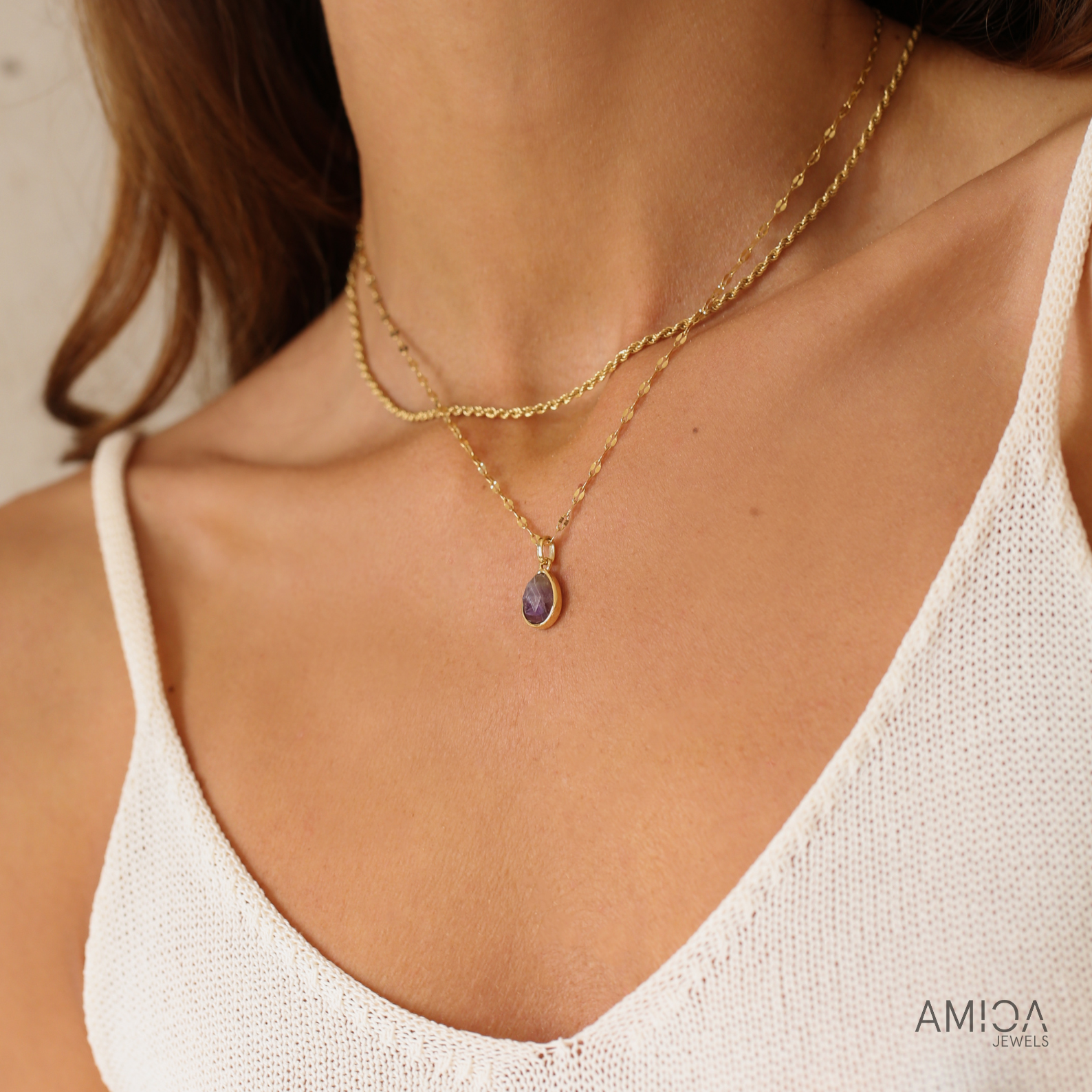 Close-up of a woman wearing a gold necklace with a purple pendant, branded 'AMICA'.