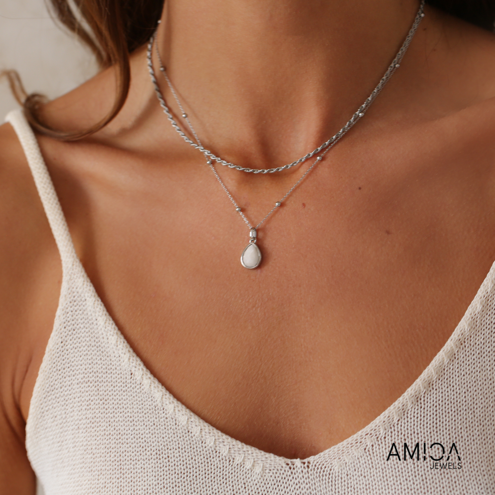 Close-up of a necklace with a pendant on a person wearing a beige top, with 'AMICA' branding.
