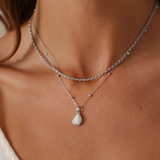 Close-up of a person wearing two silver necklaces with a pendant on a neutral background
