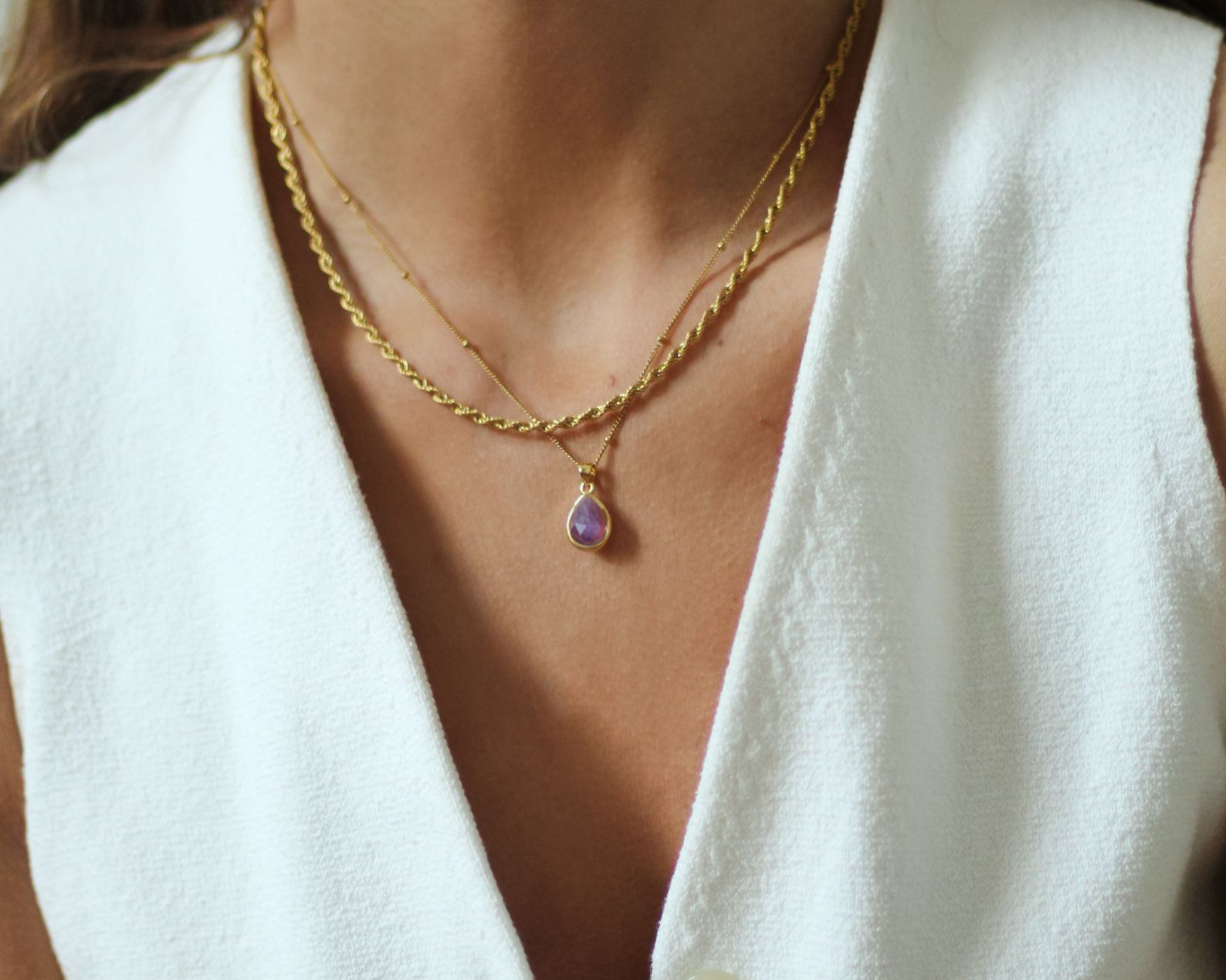 Gold necklace with a purple pendant worn by a person in a white outfit, with 'AMICA JEWELS' branding.