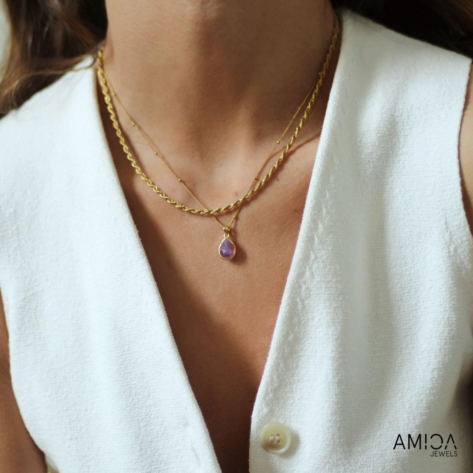 Gold necklace with a purple pendant worn by a person in a white outfit, with 'AMICA JEWELS' branding.