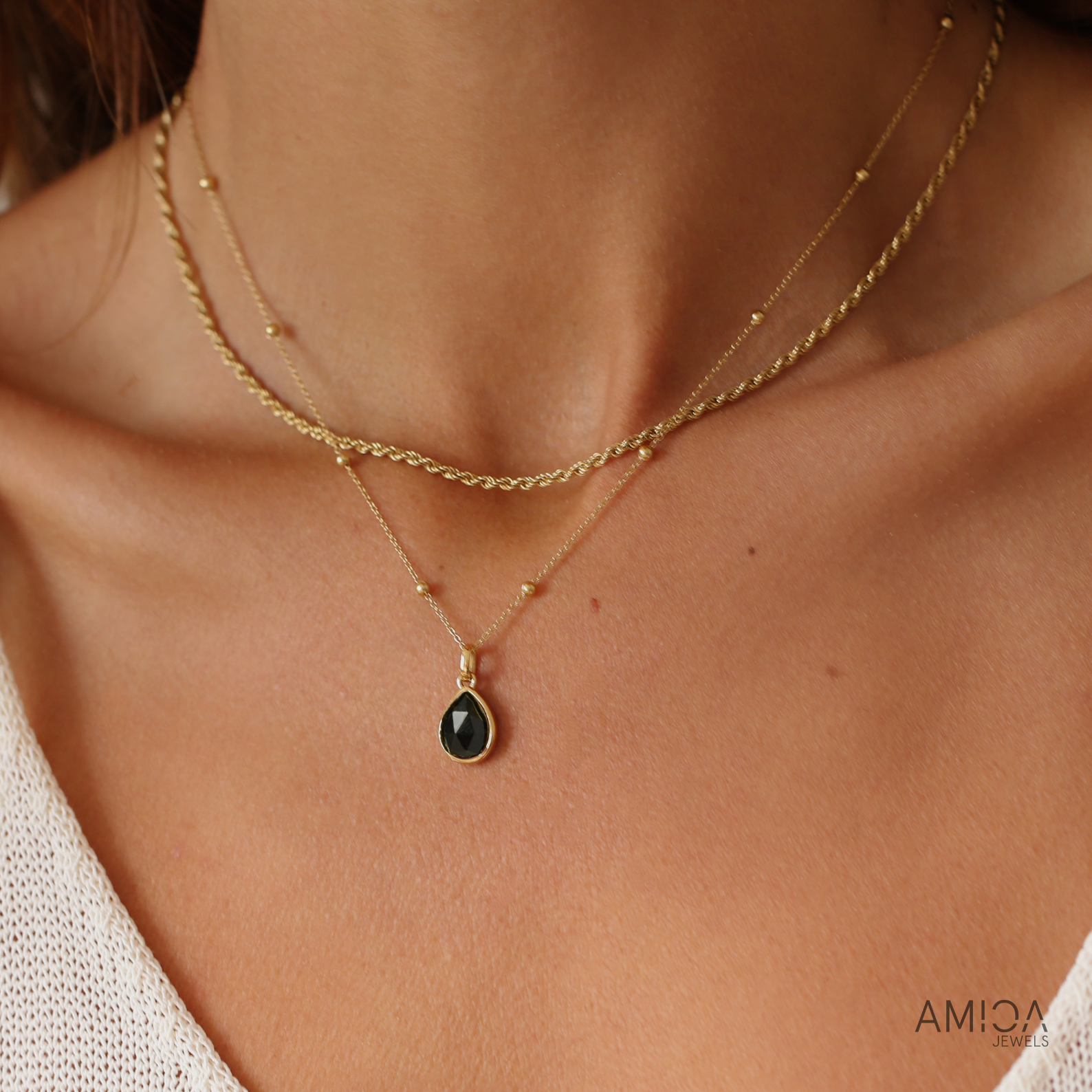 Close-up of a person wearing a gold necklace with a black pendant, brand 'AMICA' visible.