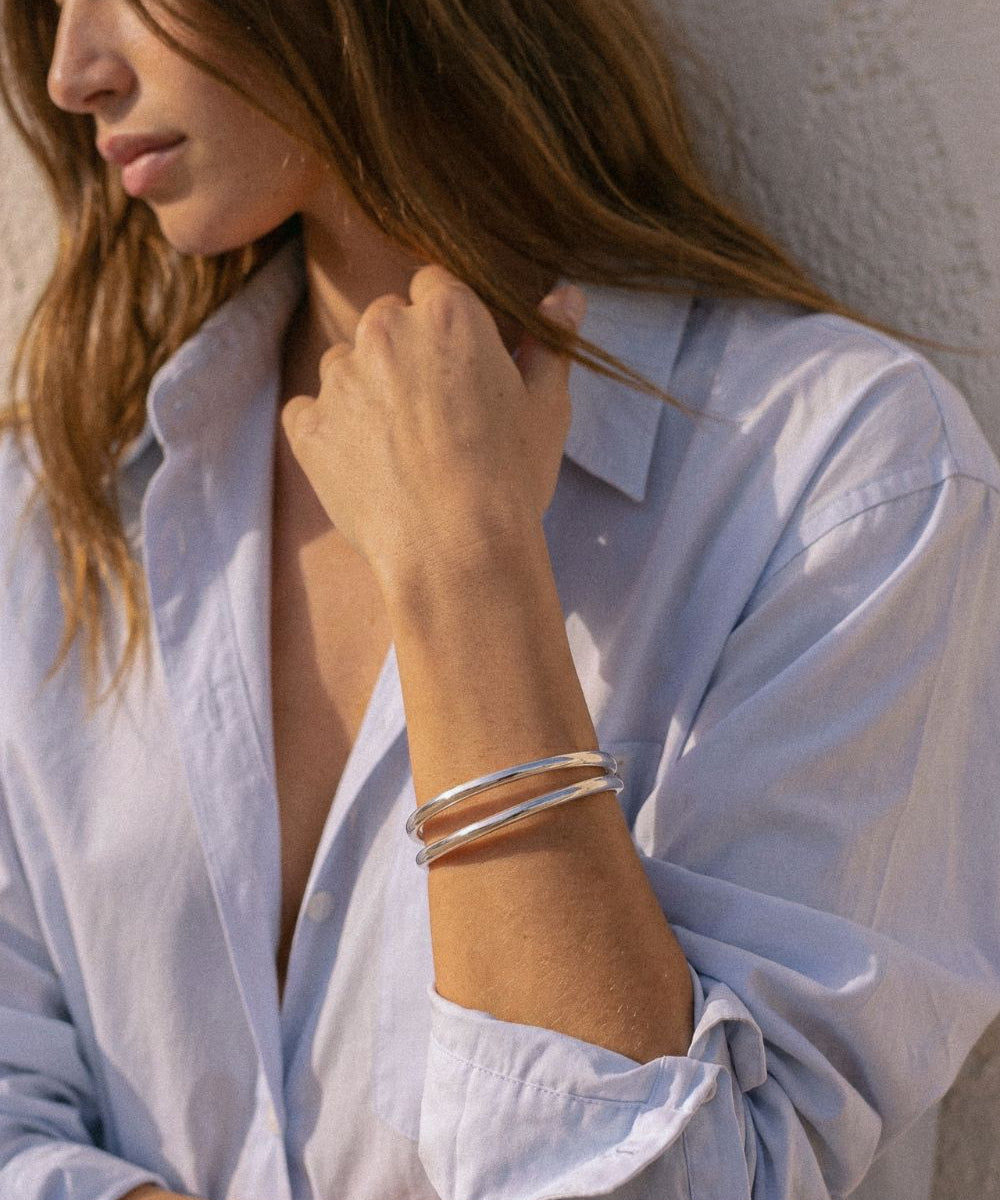 A woman wearing a stack of two thin silver bangle bracelets, styled with a light blue button-down shirt against a textured white wall.