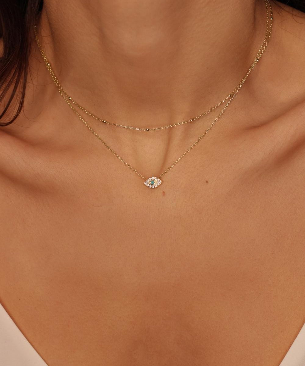 A layered necklaces featuring an evil eye charm necklace in gold and a satellite chain choker. The set includes a delicate 18k gold plated chain with an intricately designed evil eye charm pendant. Alongside the evil eye charm necklace, a satellite chain choker is also included, adding a trendy and minimalist touch. The satellite chain choker features small, evenly spaced interlinked gold beads, creating a delicate and stylish look. 