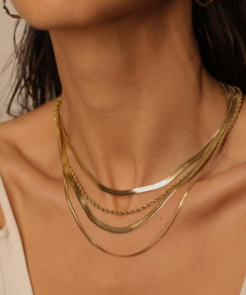 A layered set of herringbone flat chains in 18k gold plated. The set includes multiple flat chains with a distinct herringbone pattern, all crafted in a luxurious gold color. The chains are layered together, creating a stylish and modern look. The herringbone pattern adds a unique texture and visual interest to the chains, while the gold color adds a touch of glamour