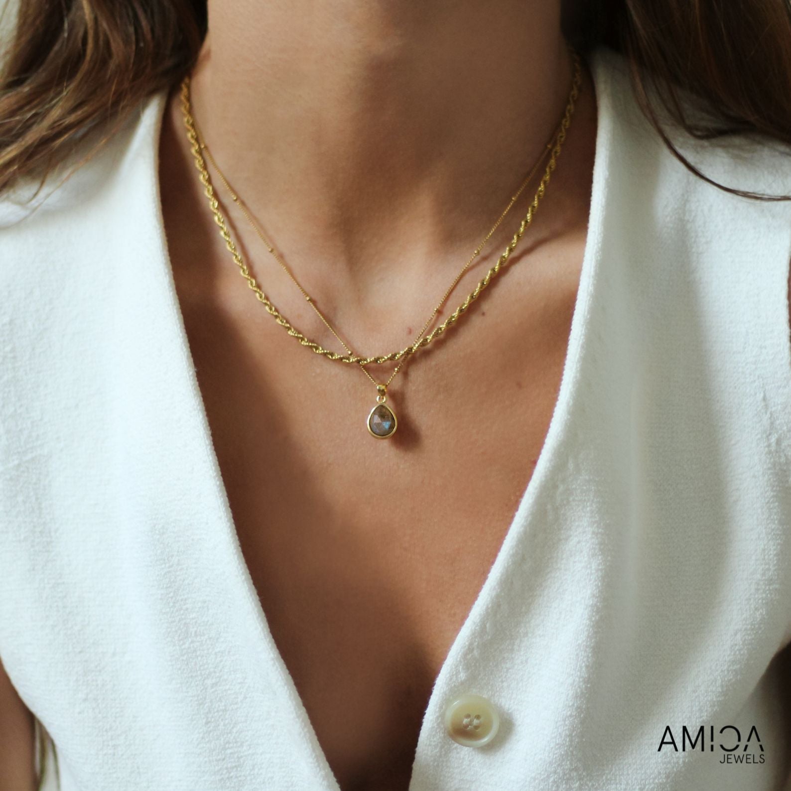 Close-up of a person wearing a gold necklace with a teardrop pendant, set against a white background. The brand 'AMICA JEWELS' is visible.