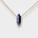Natural Lapis lazuli crystal necklace. The necklace features a pointed lapis lazuli crystal pendant with a deep blue color and golden flecks. The crystal is attached to a gold chain, creating a stunning and eye-catching piece of jewelry.