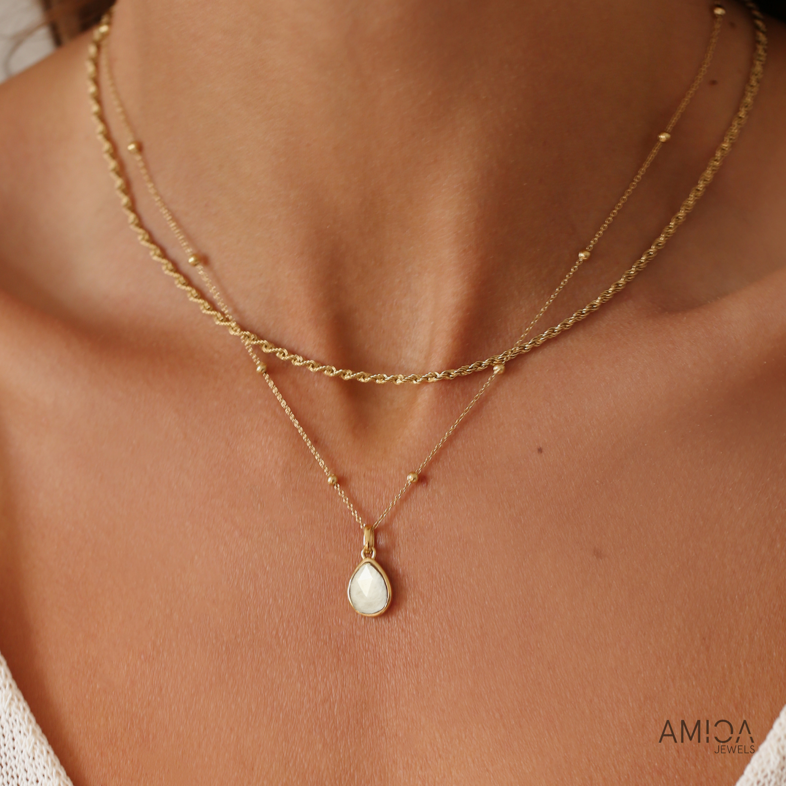 Gold necklace with a teardrop pendant on a person's neck, brand 'AMICA' visible.
