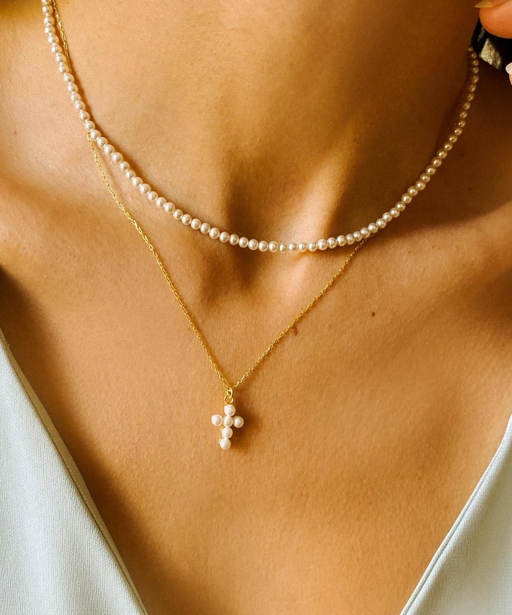 A layered pearl necklace set, featuring a pearl cross pendant necklace and a beaded pearl choker. The pearl cross pendant necklace is crafted from delicate gold-plated silver and features a cross pendant adorned with lustrous white pearls. The necklace is layered with a beaded pearl choker, which features delicate gold chains and lustrous white pearls threaded onto the chains.