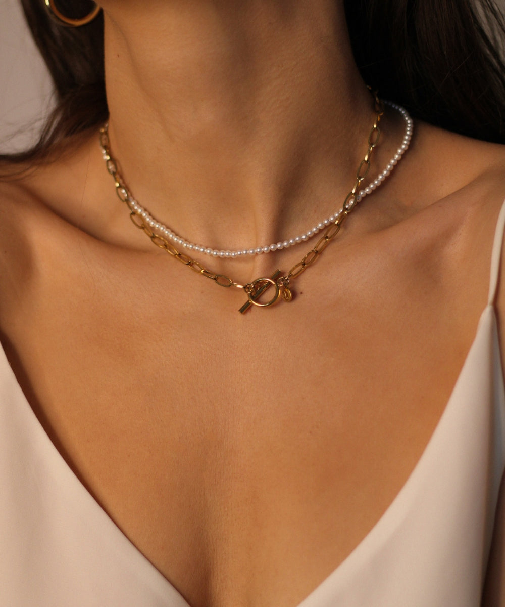 Waterproof 18k gold plated toggle choker. The set includes a sleek and minimalistic 18k gold plated toggle choker with a decorative gold ring and a bar that fits through it to secure the necklace. The choker is layered with a beaded pearl necklace, featuring lustrous white pearls strung on a delicate gold chain. The two necklaces complement each other perfectly, adding a touch of elegance and sophistication to the model's outfit