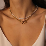 Waterproof 18k gold plated toggle choker. The set includes a sleek and minimalistic 18k gold plated toggle choker with a decorative gold ring and a bar that fits through it to secure the necklace. The choker is layered with a beaded pearl necklace, featuring lustrous white pearls strung on a delicate gold chain. The two necklaces complement each other perfectly, adding a touch of elegance and sophistication to the model's outfit