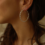 a pair of silver hoop earrings, featuring a twisted design. The hoops are large in size, yet lightweight, making them comfortable to wear. The silver has a polished finish, reflecting light and enhancing their shine. These striking earrings add a bold and elegant touch to any outfit.