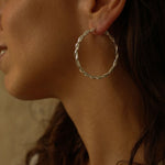 a pair of silver hoop earrings, featuring a twisted design. The hoops are large in size, yet lightweight, making them comfortable to wear. The silver has a polished finish, reflecting light and enhancing their shine. These striking earrings add a bold and elegant touch to any outfit.