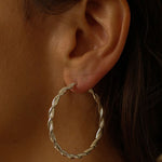 a pair of silver hoop earrings, featuring a twisted design. The hoops are large in size, yet lightweight, making them comfortable to wear. The silver has a polished finish, reflecting light and enhancing their shine. These striking earrings add a bold and elegant touch to any outfit.