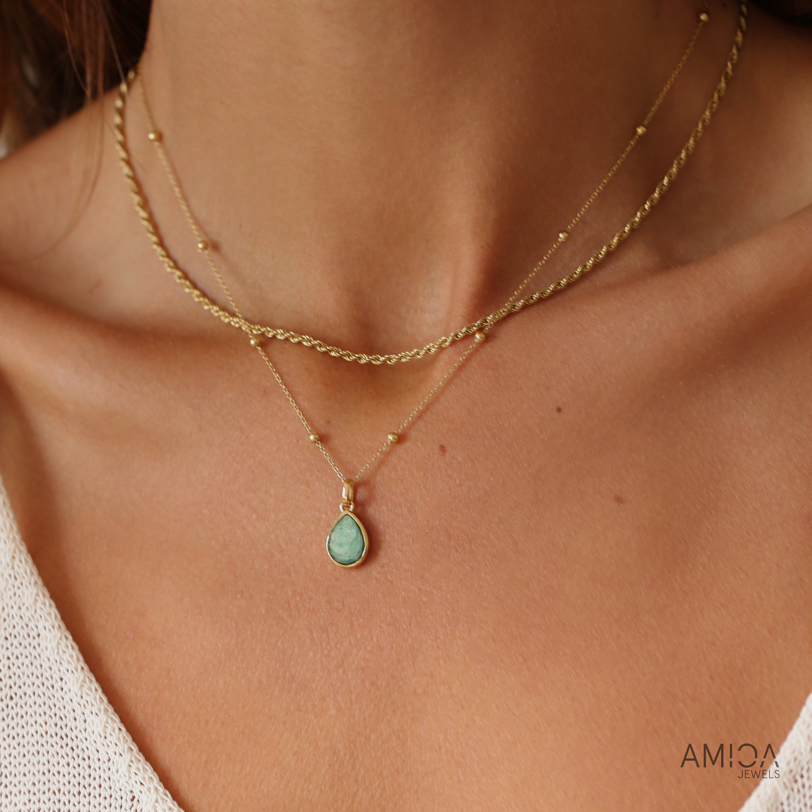 Close-up of a person wearing a gold necklace with a green teardrop pendant, brand 'AMICA' visible.