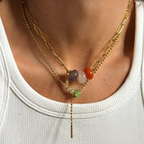 A close-up of a person's neck wearing a layered gold necklace with a paperclip chain, a beaded chain, and a central drop chain featuring colorful gemstone beads.
