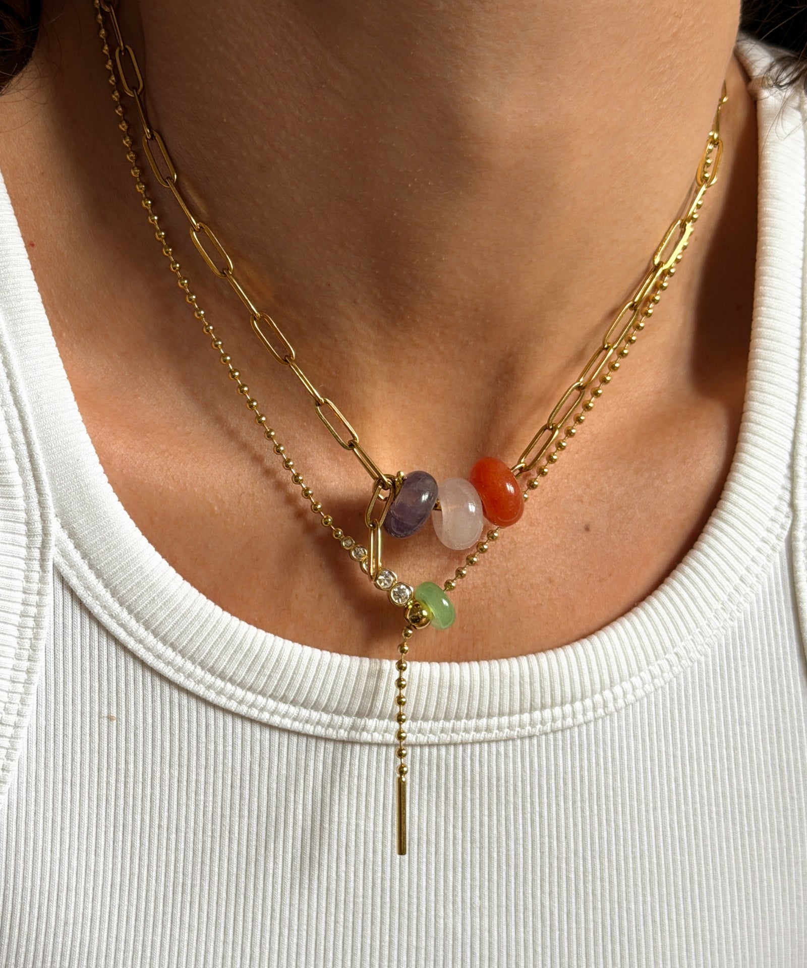 A close-up of a person's neck wearing a layered gold necklace with a paperclip chain, a beaded chain, and a central drop chain featuring colorful gemstone beads.