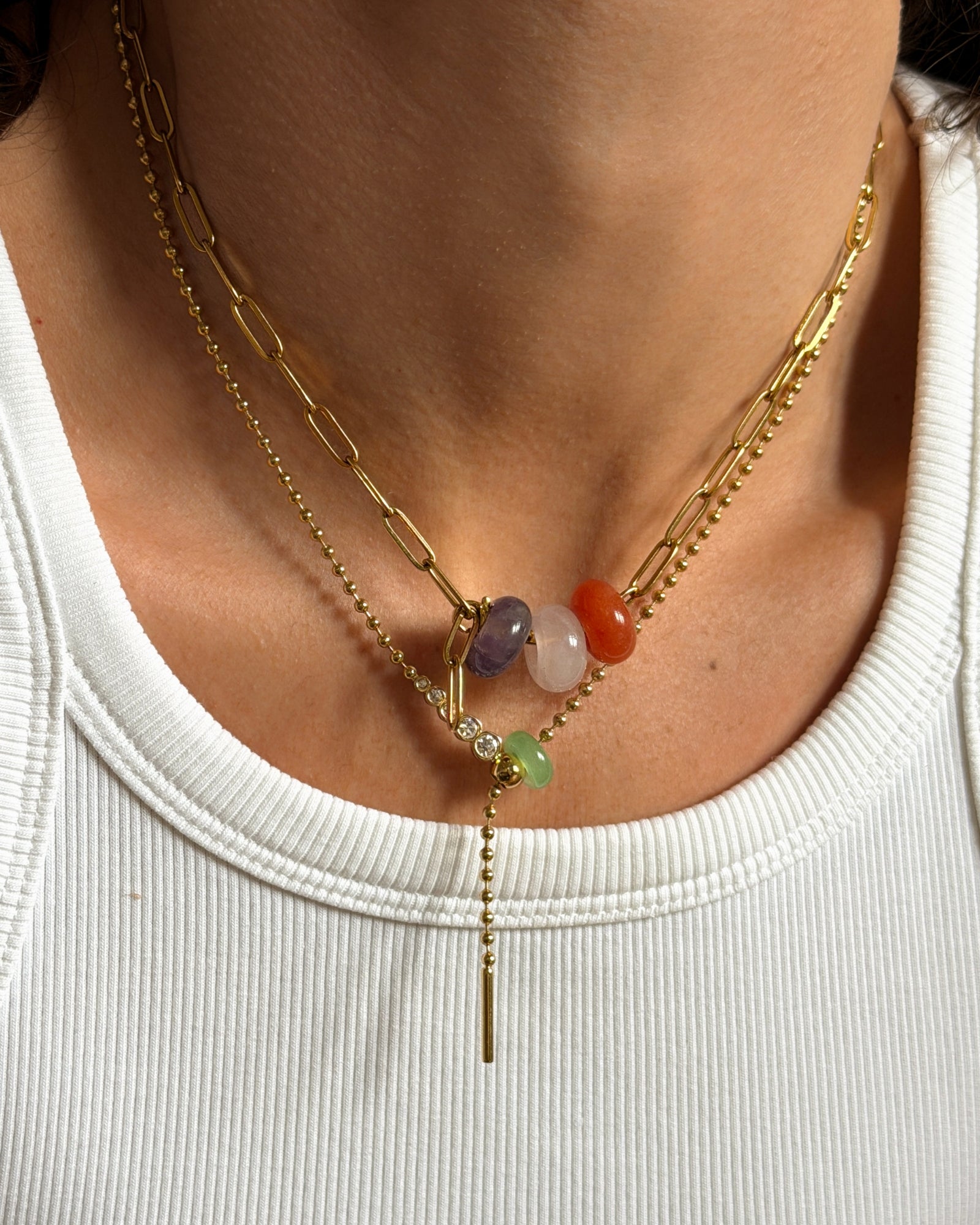 A close-up of a person's neck wearing a layered gold necklace with a paperclip chain, a beaded chain, and a central drop chain featuring colorful gemstone beads.