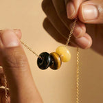 Close-up of a gold chain necklace with three round beads: black obsidian,  tigers eye and yellow jade. Hands are holding the necklace against a warm beige background.