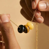 Close-up of a gold chain necklace with three round beads: black obsidian,  tigers eye and yellow jade. Hands are holding the necklace against a warm beige background.