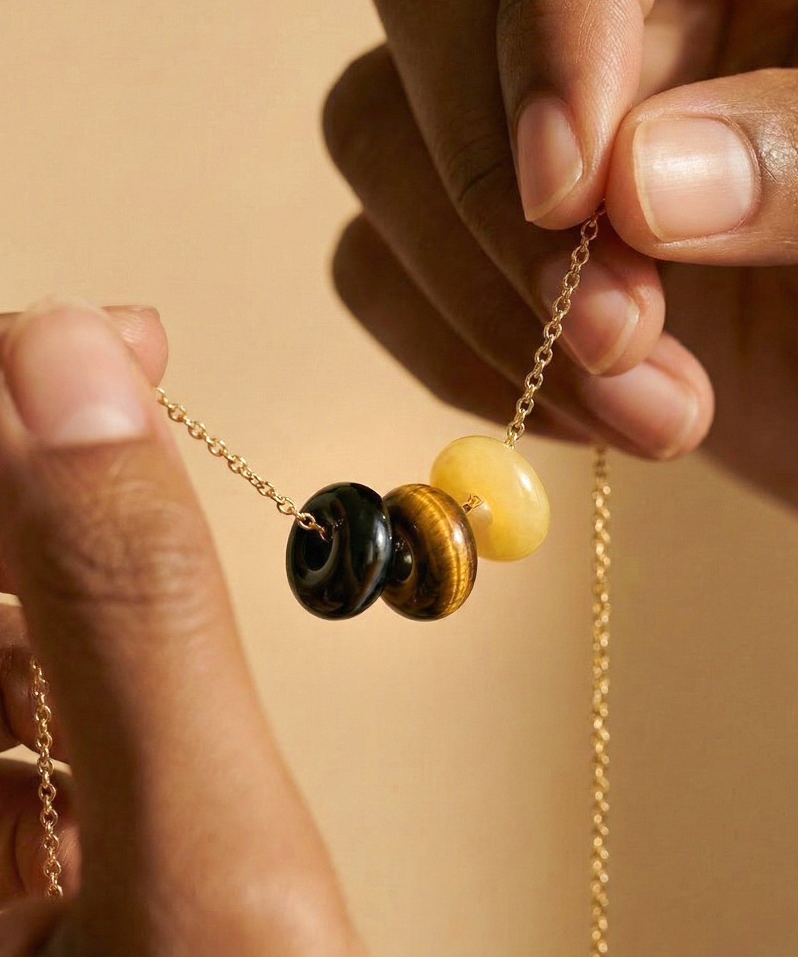 Close-up of a gold chain necklace with three round beads: black obsidian,  tigers eye and yellow jade. Hands are holding the necklace against a warm beige background.