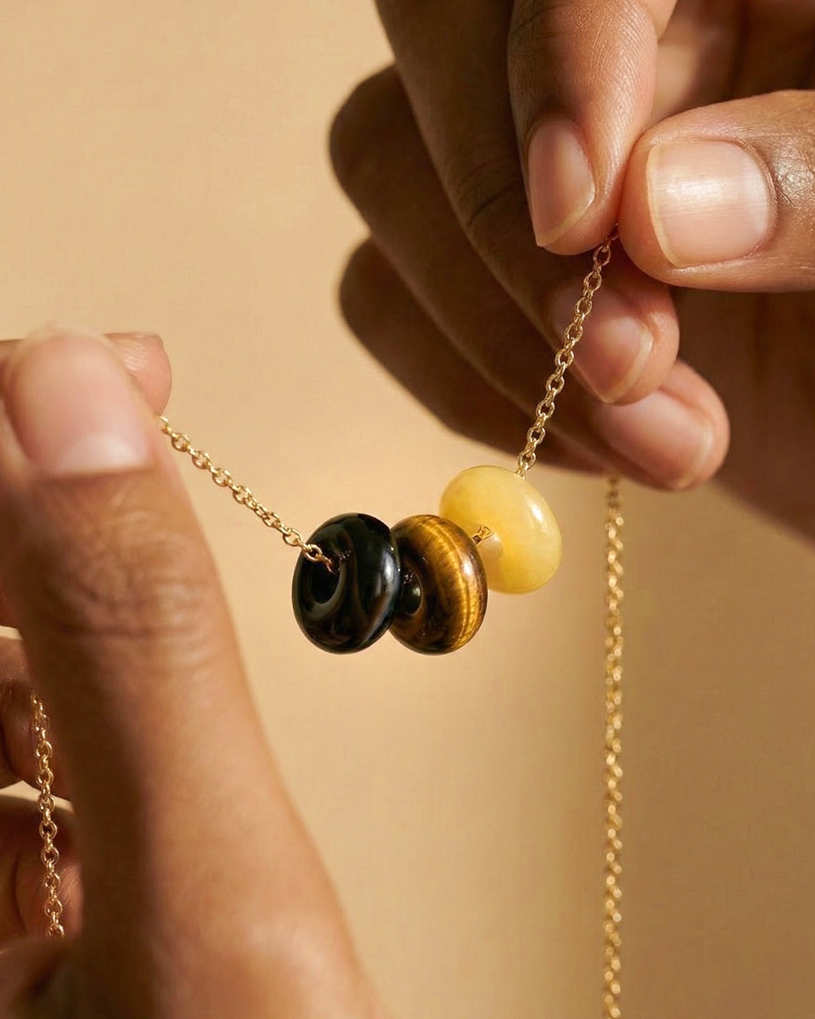 Close-up of a gold chain necklace with three round beads: black obsidian,  tigers eye and yellow jade. Hands are holding the necklace against a warm beige background.