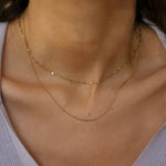 A close-up of a model's neck and upper chest wearing two delicate gold chain necklaces layered over a light lavender ribbed top.