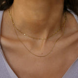 A close-up of a model's neck and upper chest wearing two delicate gold chain necklaces layered over a light lavender ribbed top.