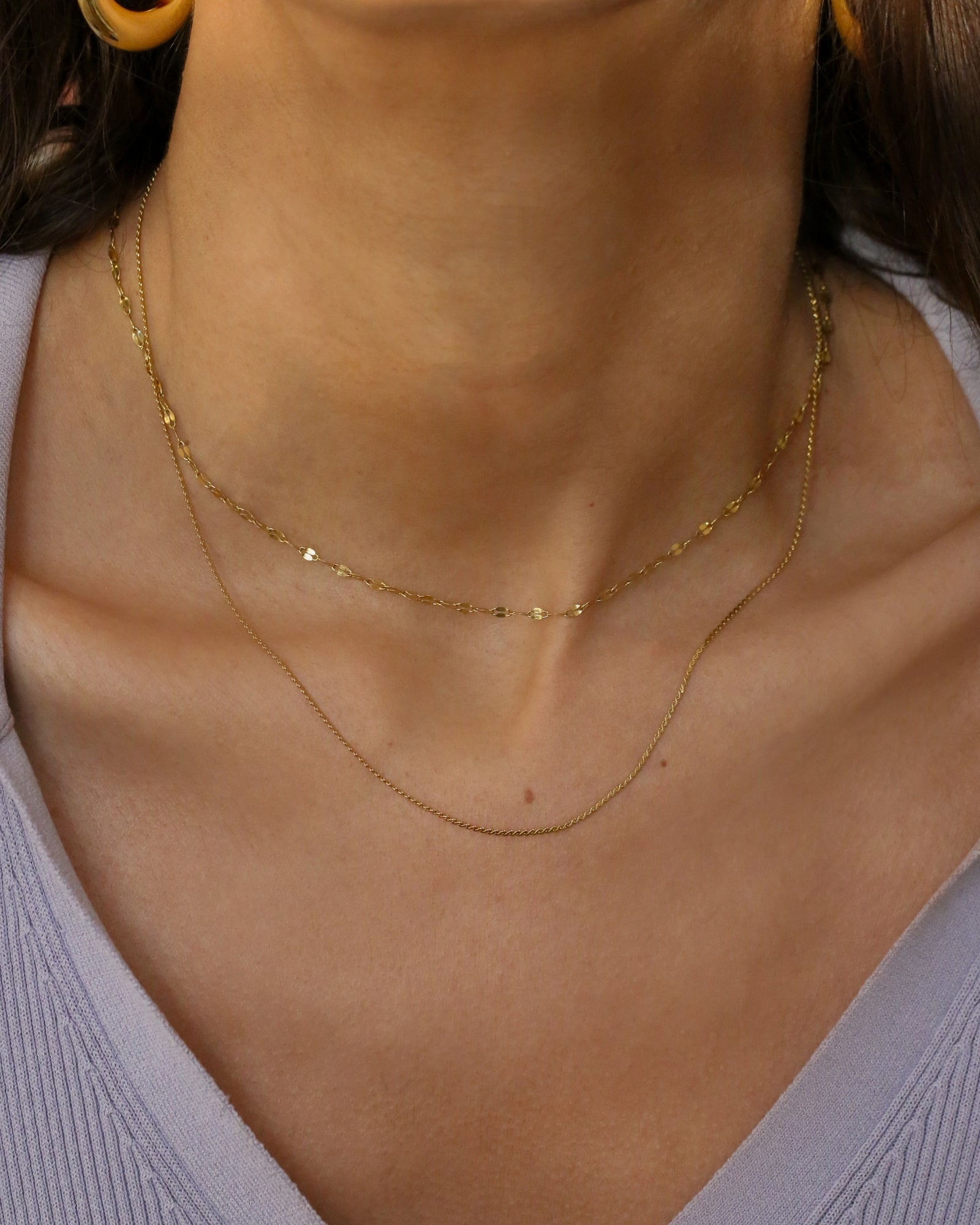 A close-up of a model's neck and upper chest wearing two delicate gold chain necklaces layered over a light lavender ribbed top.