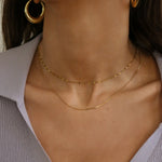 Close-up of a model's neck and upper chest wearing two delicate gold necklaces layered with matching gold hoop earrings, paired with a light lavender ribbed top.