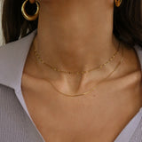 Close-up of a model's neck and upper chest wearing two delicate gold necklaces layered with matching gold hoop earrings, paired with a light lavender ribbed top.