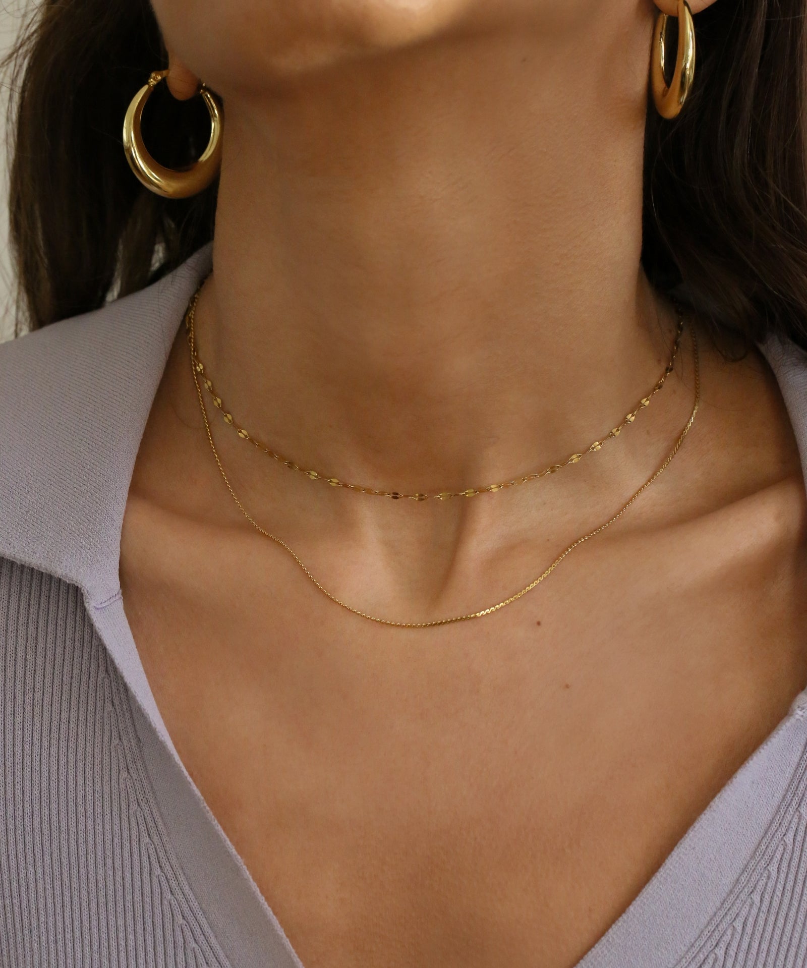 Close-up of a model's neck and upper chest wearing two delicate gold necklaces layered with matching gold hoop earrings, paired with a light lavender ribbed top.