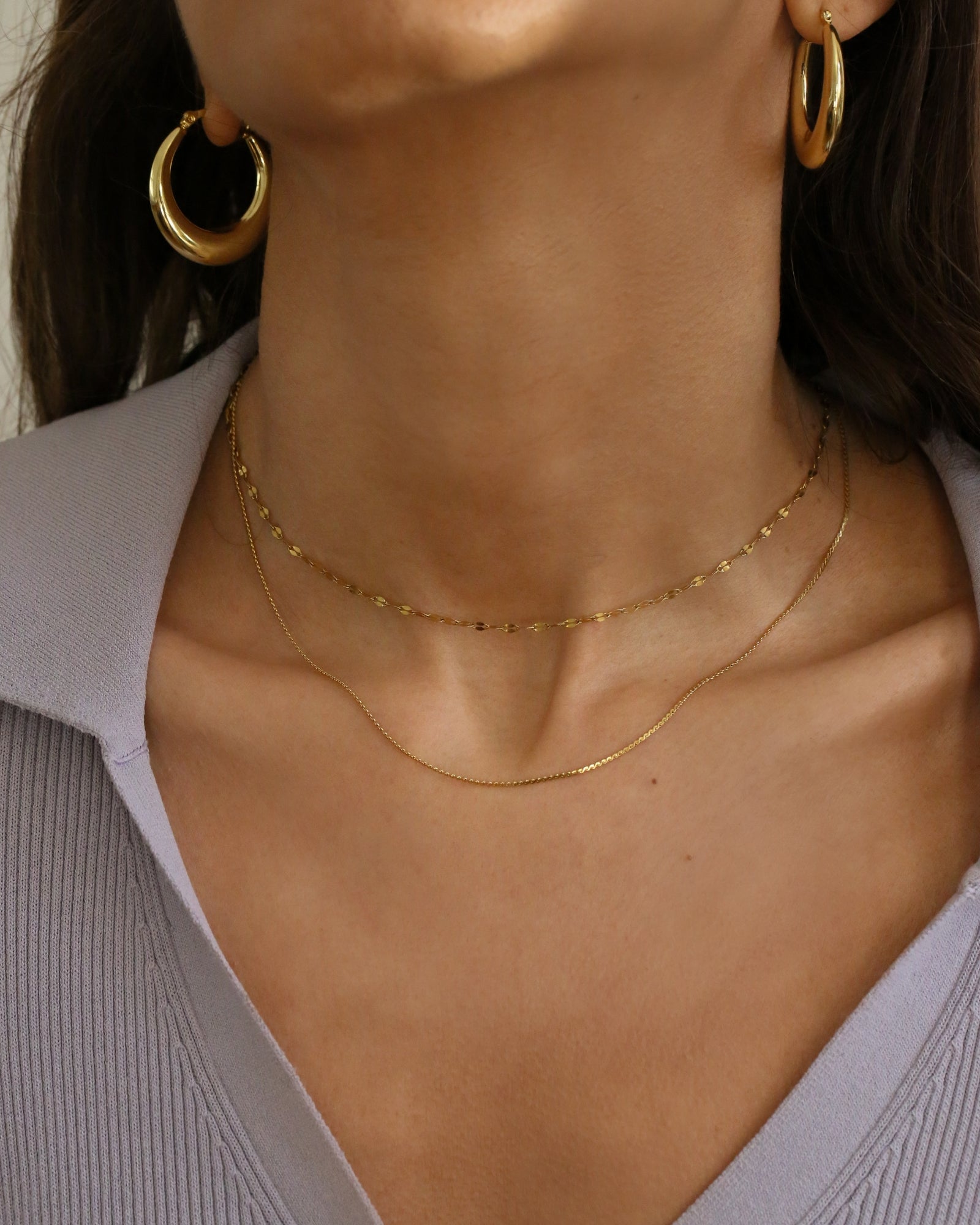 Close-up of a model's neck and upper chest wearing two delicate gold necklaces layered with matching gold hoop earrings, paired with a light lavender ribbed top.